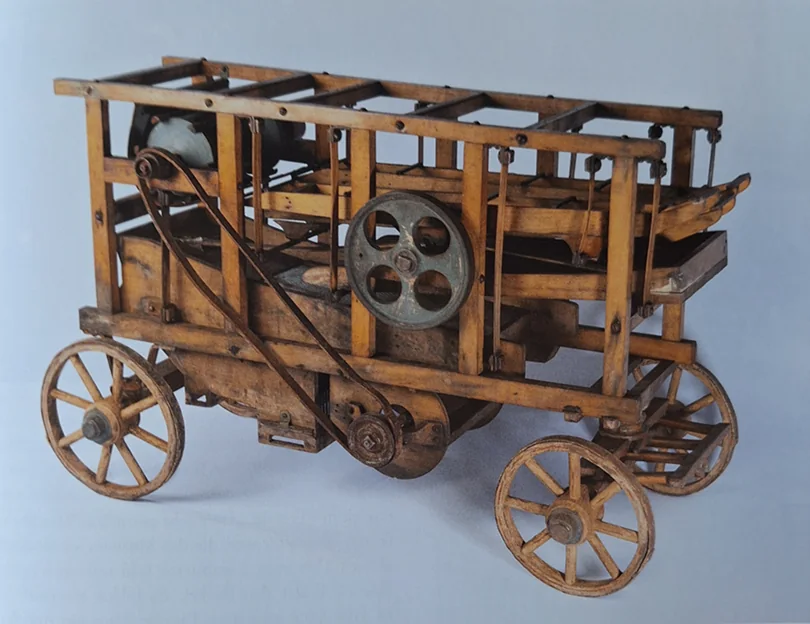 Hölzernes Modell einer Dreschmaschine auf vier Speichenrädern mit Metallschwungrad und Antriebsriemen.