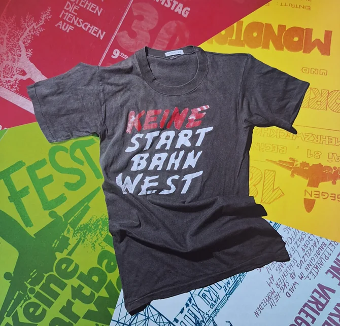 Dunkelgraues T-Shirt mit handgemaltem Schriftzug „Keine Startbahn West“, liegend auf bunten Protestplakaten.